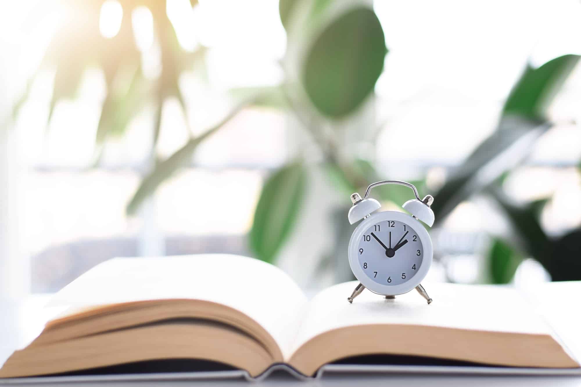 Clock on top of an open book with blurry green houseplants in the background. Reading time